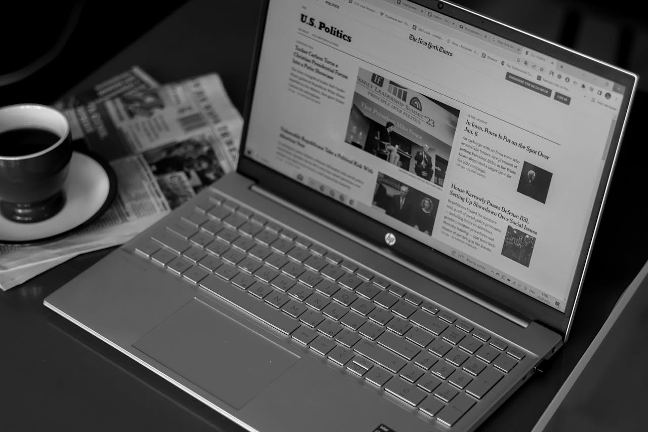gallery-1 Black and white image of a laptop displaying news articles, accompanied by a cup of coffee and newspapers.